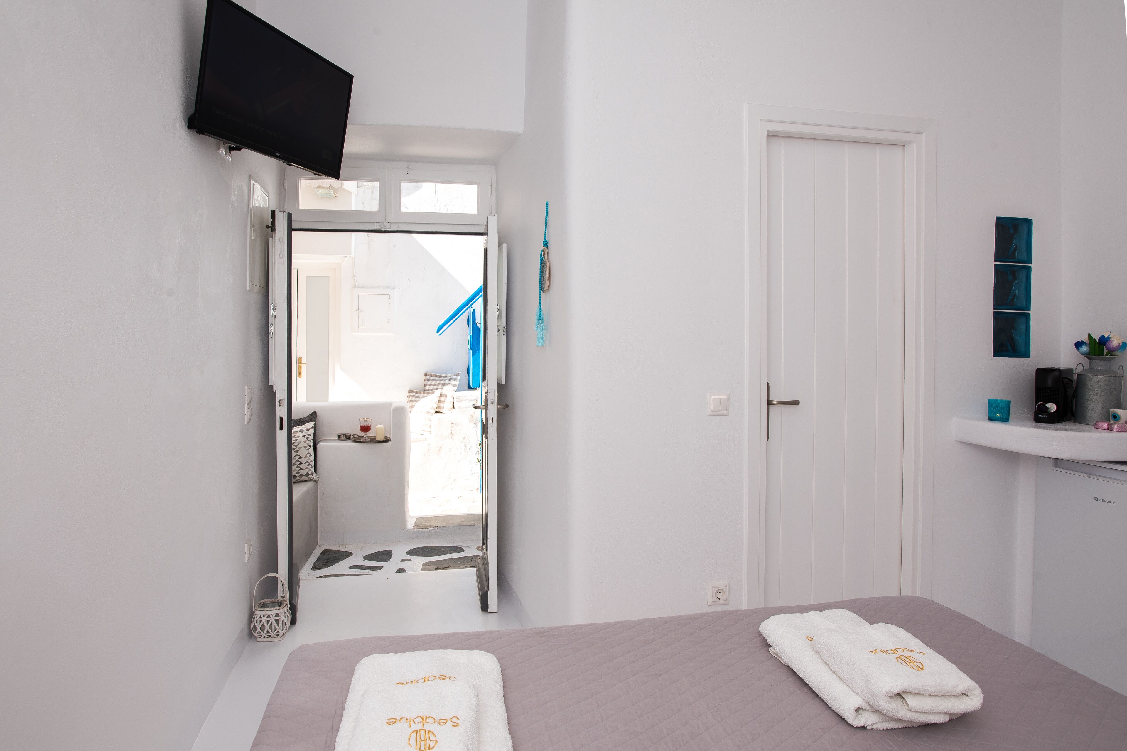 Habitación de hotel con cama, televisor montado en la pared, baño visible a través de una puerta abierta y un estante pequeño con artículos de tocador.