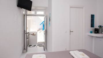 Habitación de hotel con cama, televisor montado en la pared, baño visible a través de una puerta abierta y un estante pequeño con artículos de tocador.