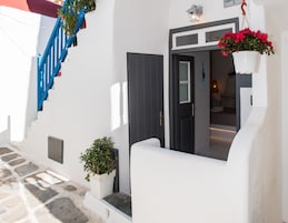 Una calle estrecha con un edificio blanco, una escalera azul y una puerta que da a una habitación con una cama y una planta colgante.