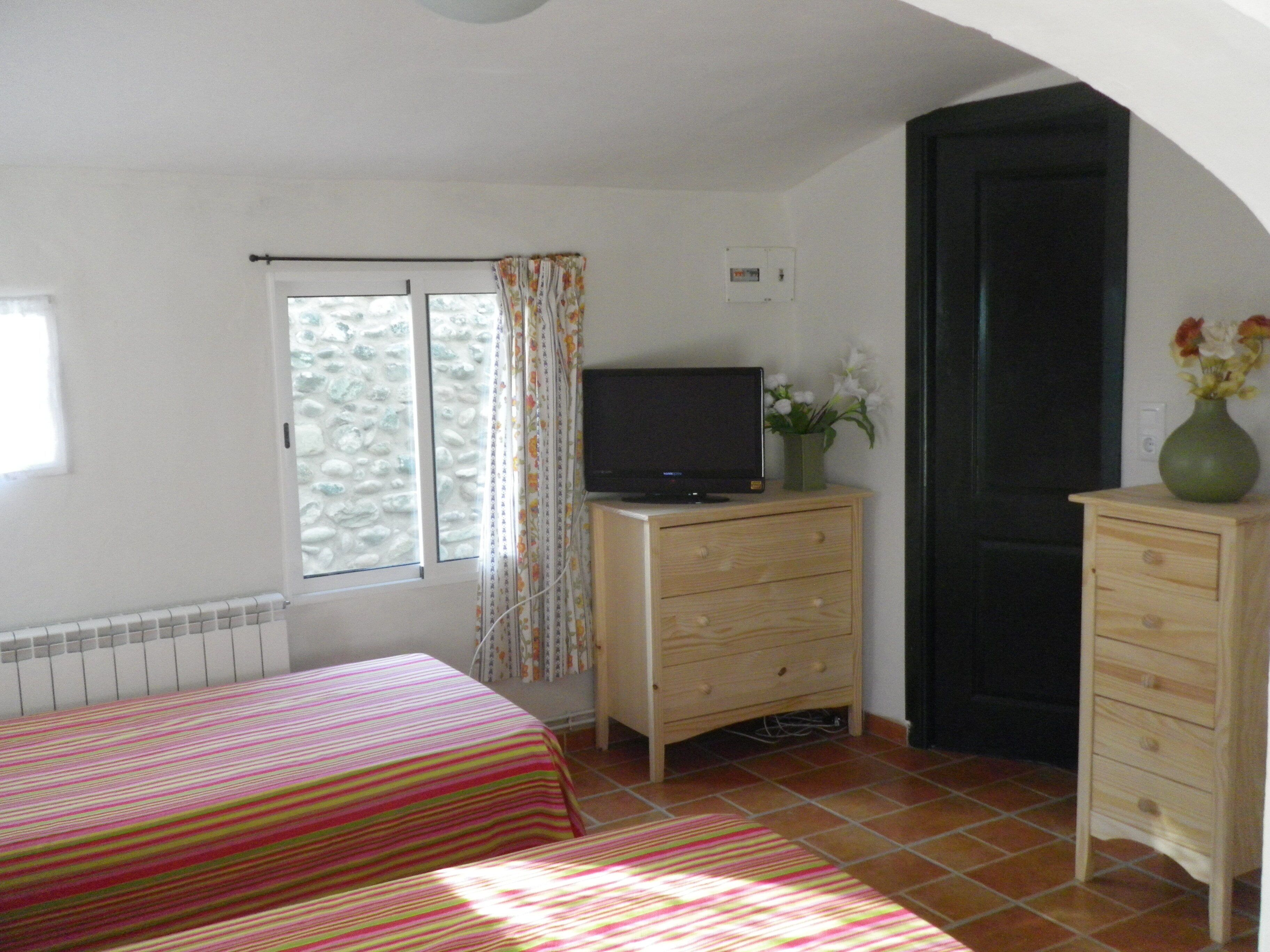 Een slaapkamer met twee bedden, een televisie op een houten dressoir, een raam met gordijnen en een deur.