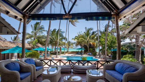 A covered outdoor seating area with wicker furniture, a pool, and palm trees in the background. - Diani Blue (Diani Beach)