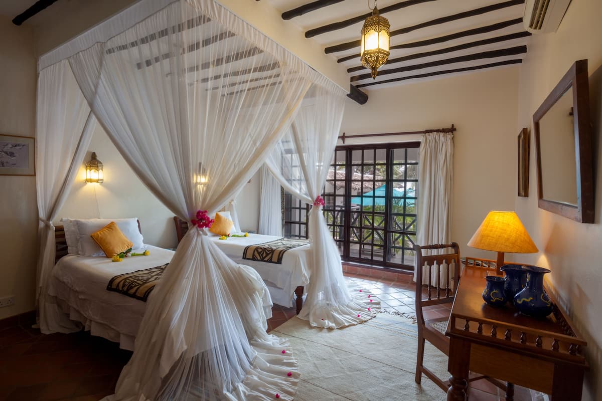 A bedroom with a canopy bed, a wooden table with a lamp, and a view through a window.