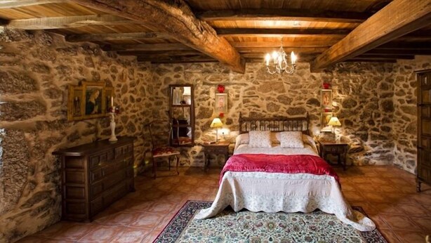 Un dormitorio rústico con paredes de piedra, vigas de madera, una cama grande y una araña.
