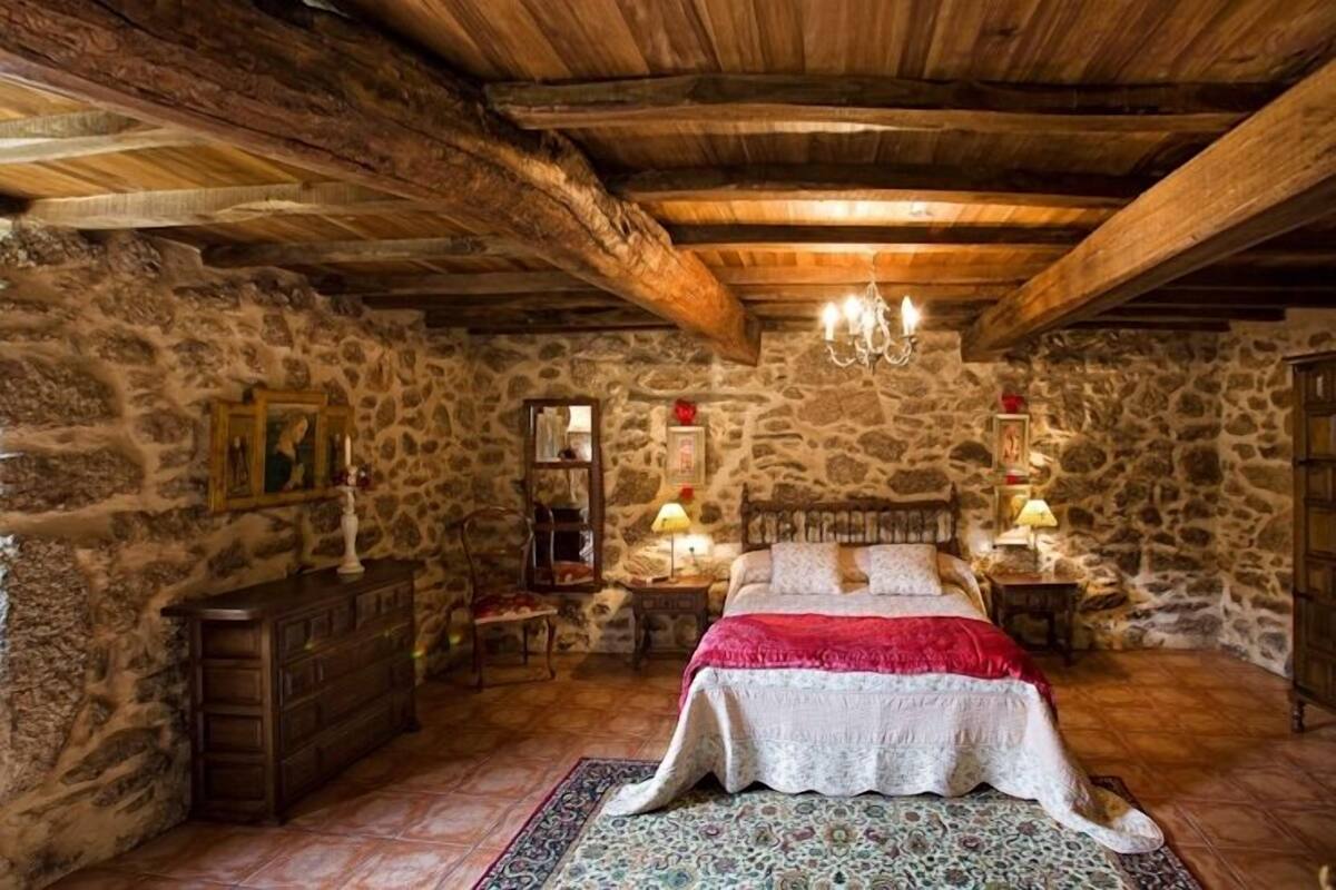 Un dormitorio rústico con paredes de piedra, vigas de madera, una cama grande y una araña.