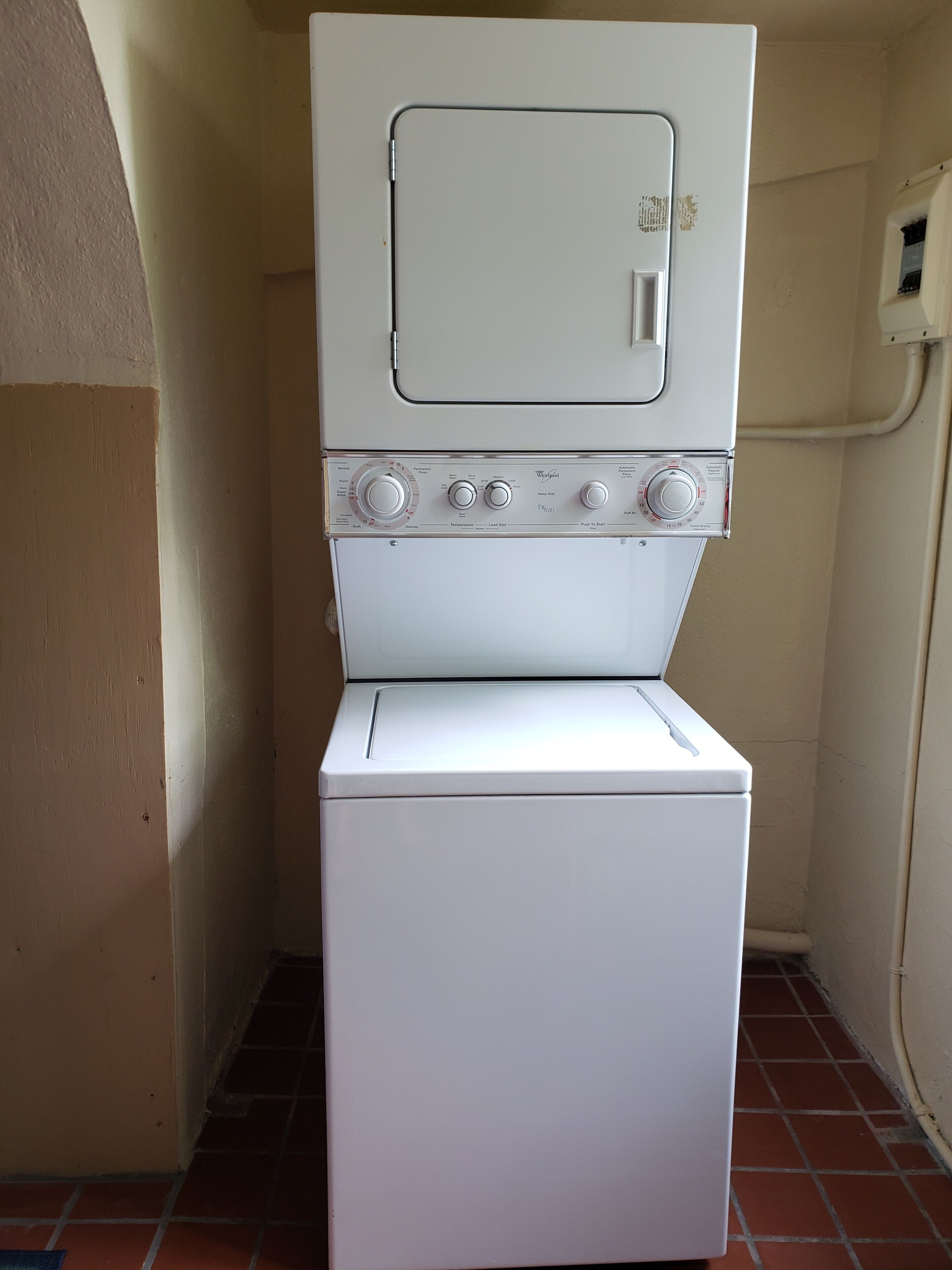 Una lavatrice e asciugatrice bianche con manopole di controllo e un'anta, installate in un angolo con pavimento piastrellato e pareti color beige.