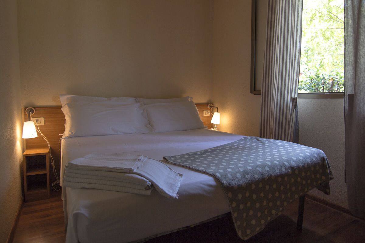 Een opgemaakt bed met witte lakens, een bedlampje en een raam met gordijnen.