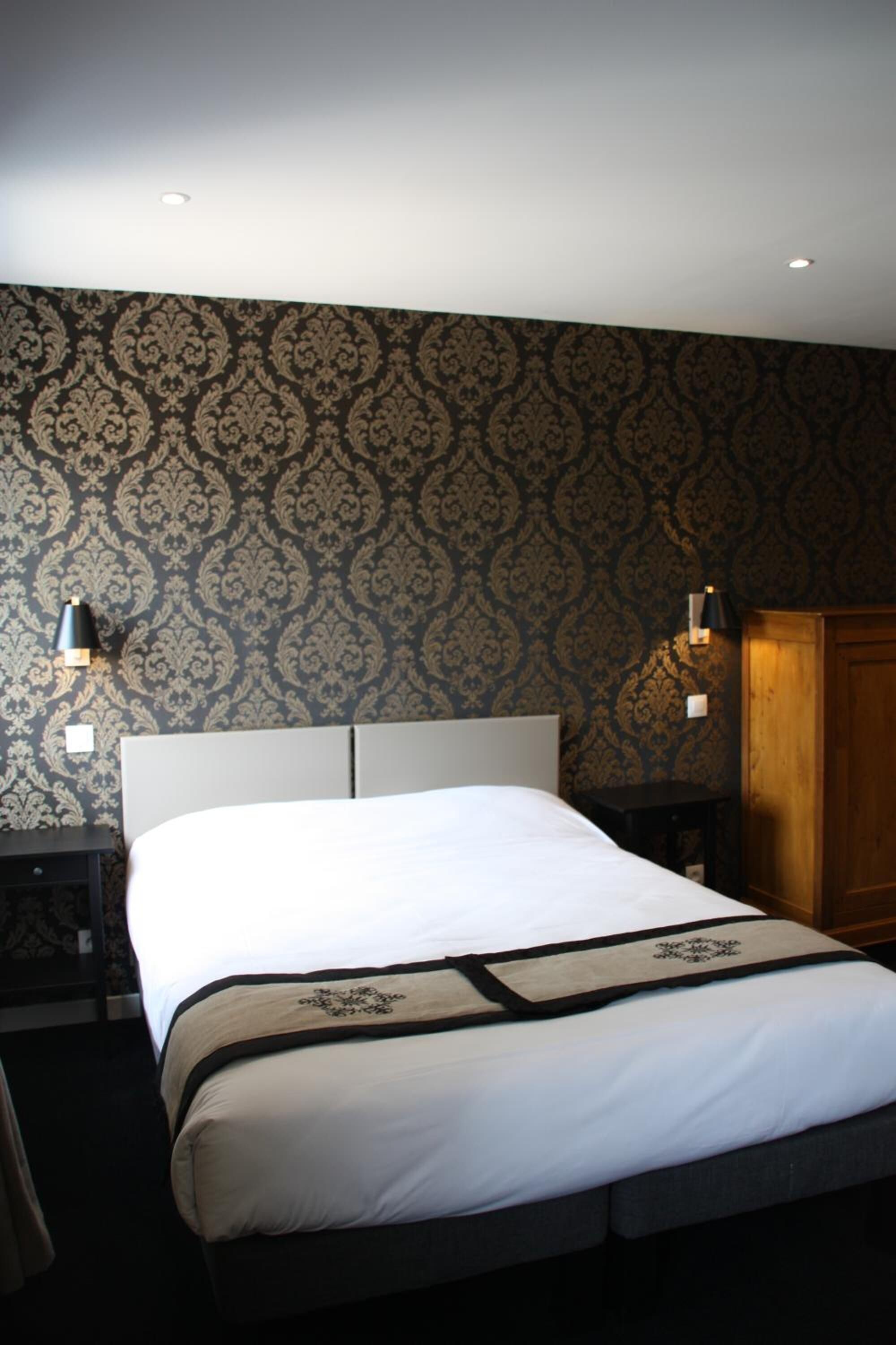 Ein Hotelzimmer mit einem großen Bett, zwei Nachttischen, einem Holzschrank und ornamentalem Wandpapier.