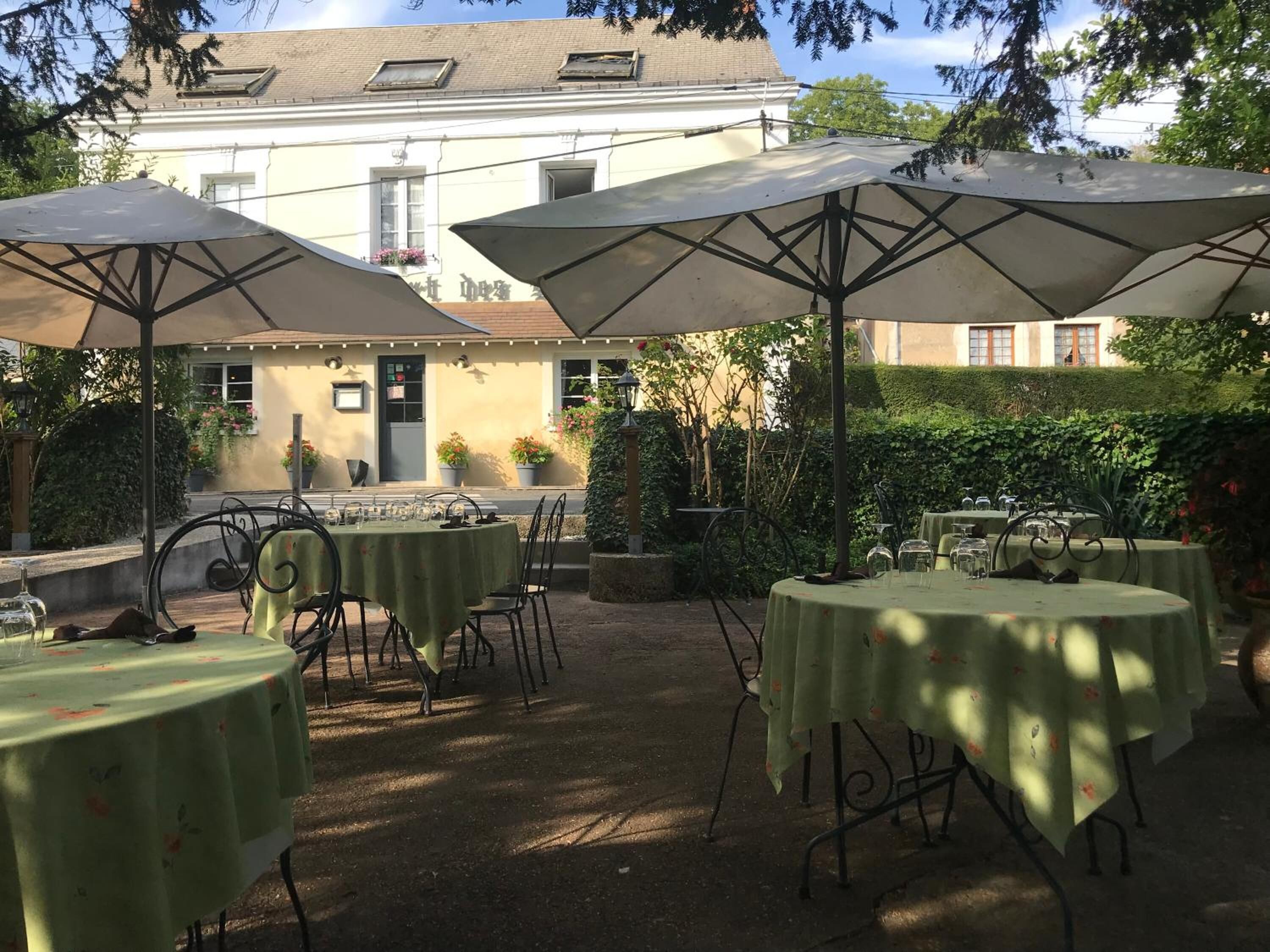 An outdoor patio with tables and umbrellas, surrounded by greenery and a building in the background.