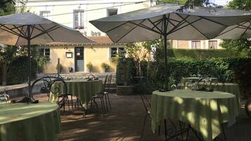 An outdoor patio with tables and umbrellas, surrounded by greenery and a building in the background.