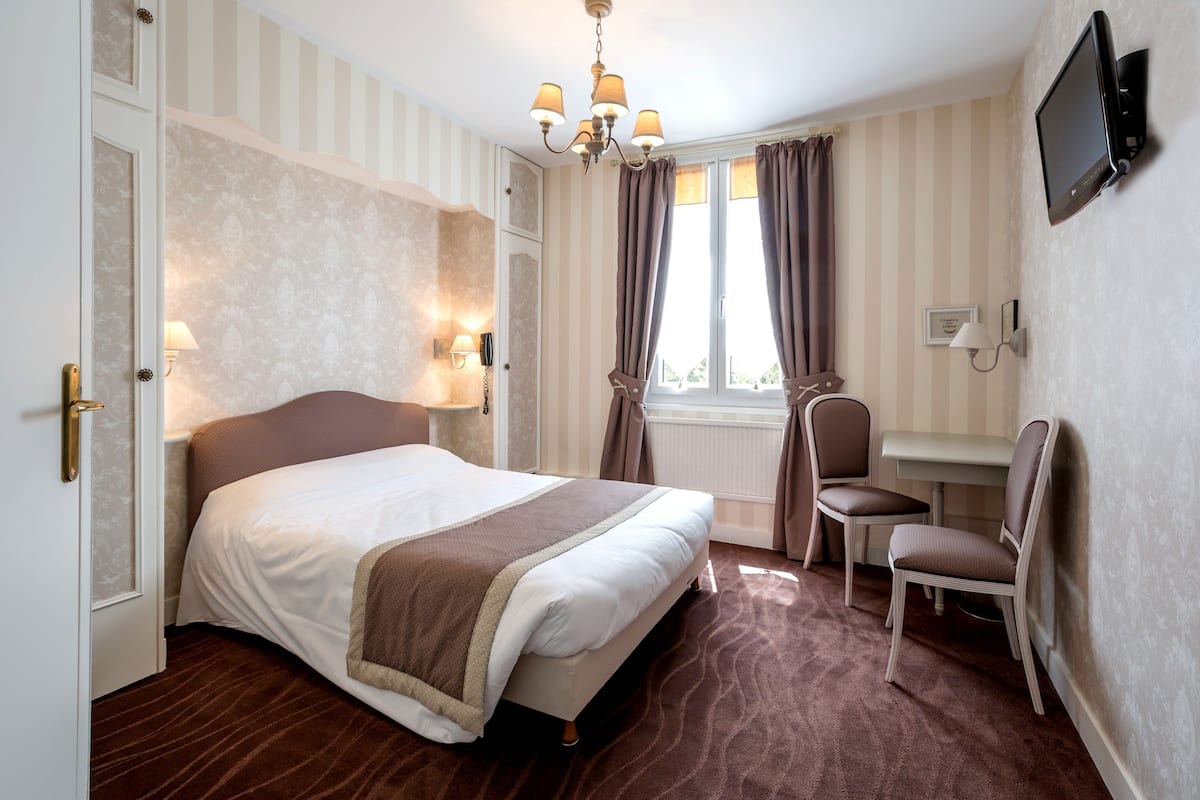 A hotel room with a large bed, a desk with a chair, a television mounted on the wall, and a window with curtains.