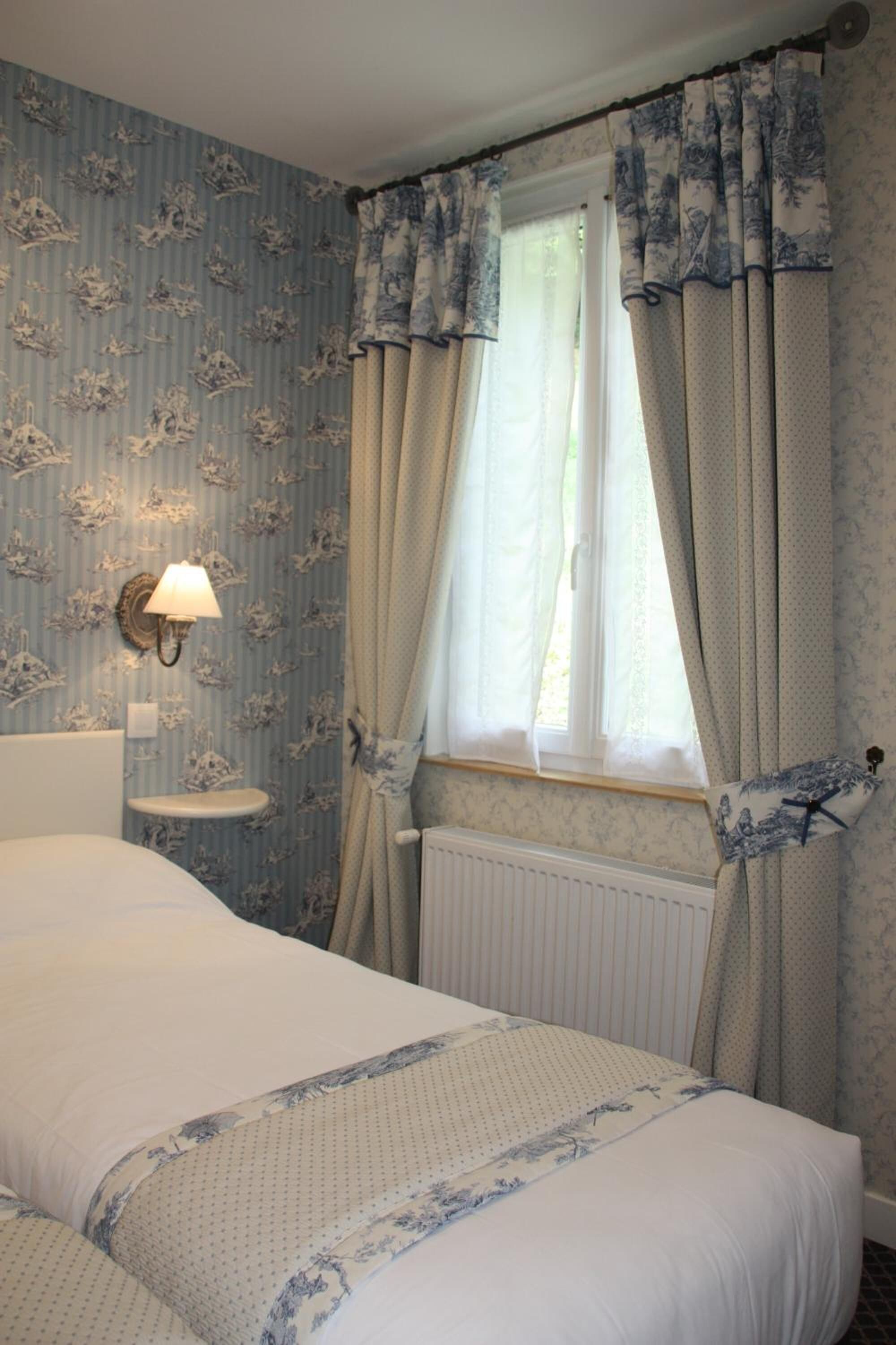 A bedroom with a bed, a nightstand, a wall-mounted lamp, and a window with curtains.