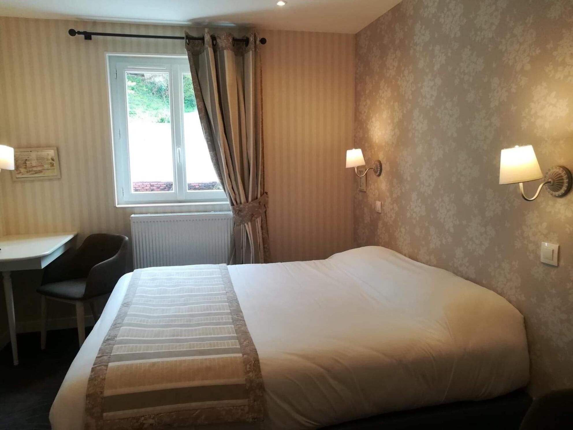 Habitación de hotel con una cama grande, un escritorio, dos lámparas de pared, una ventana con cortinas y un radiador.