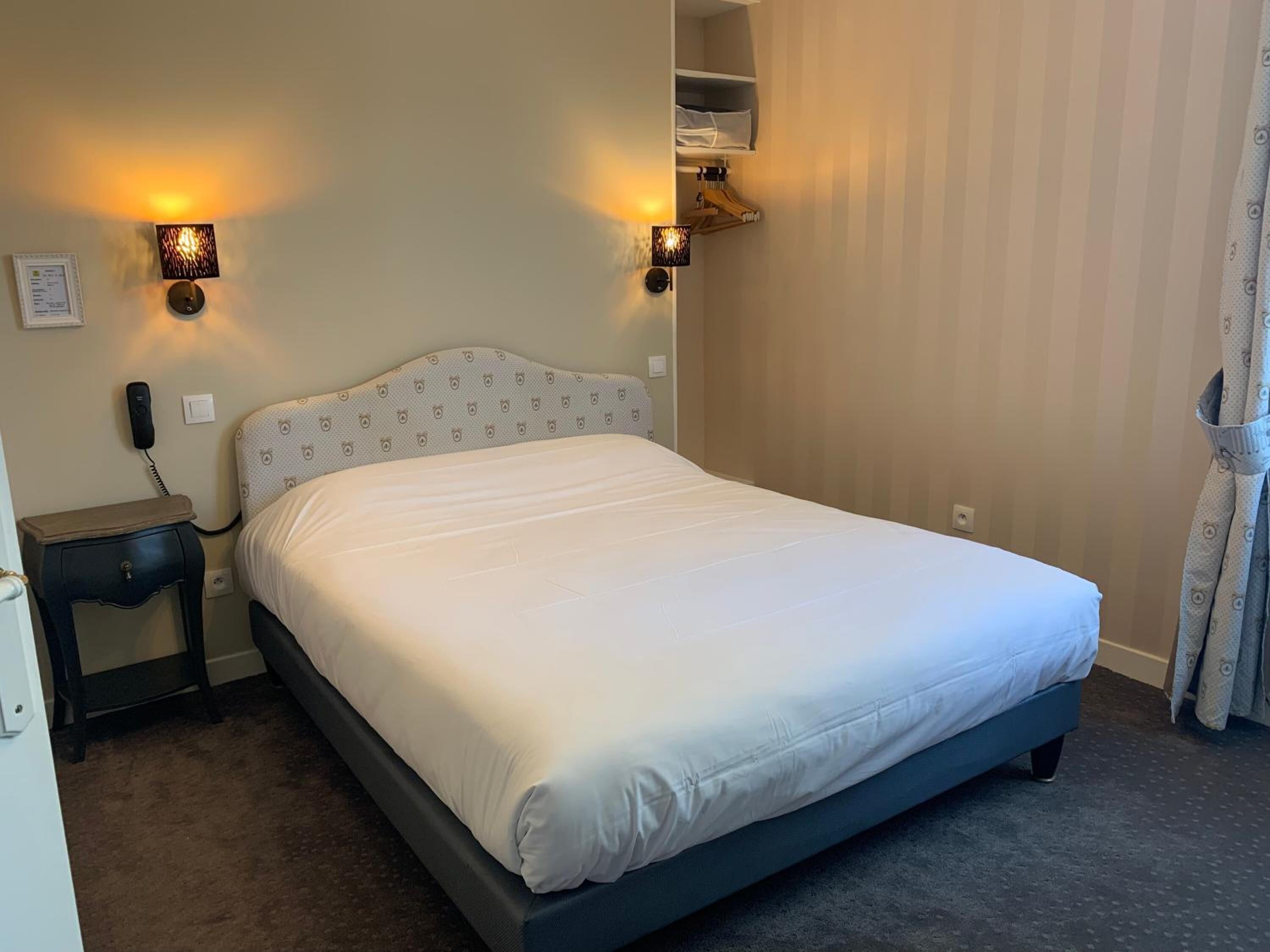 A hotel room with a bed, bedside tables, a telephone, wall-mounted lamps, and a hanging coat rack.