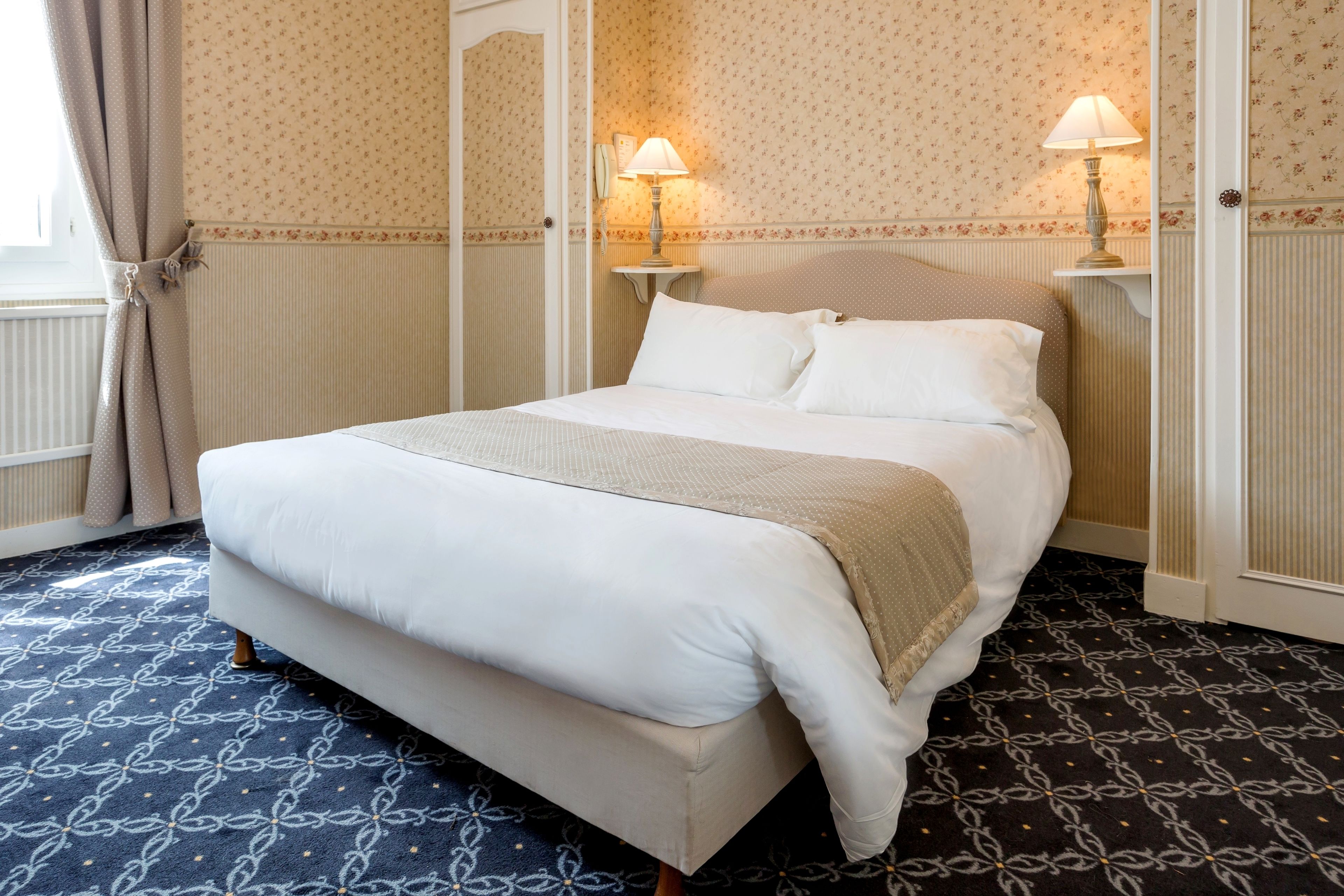 Un lit bien fait, recouvert d’une couverture beige, avec deux lampes de chevet et un tapis à motifs.