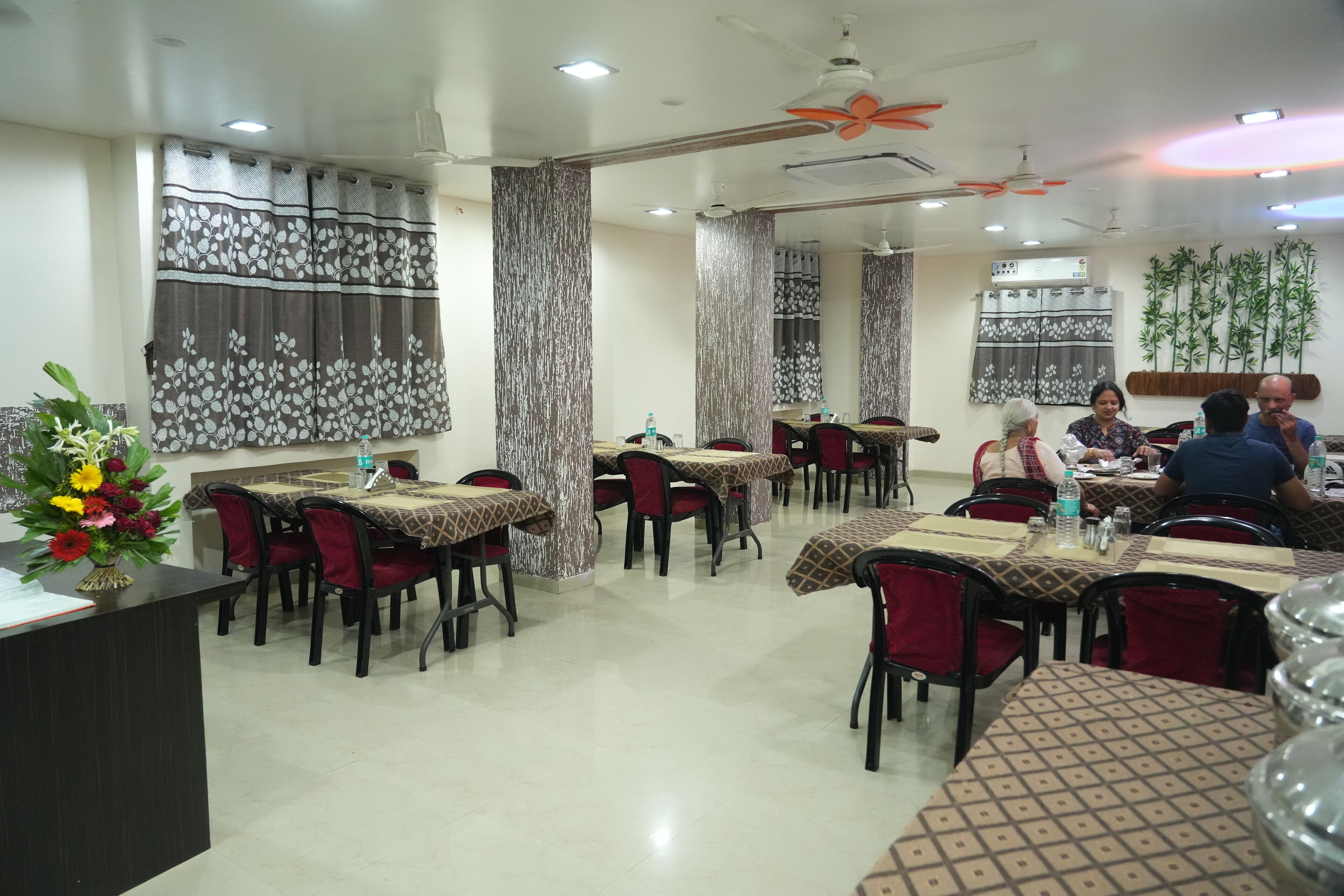 A restaurant with multiple tables and chairs, a reception desk, and decorative plants.