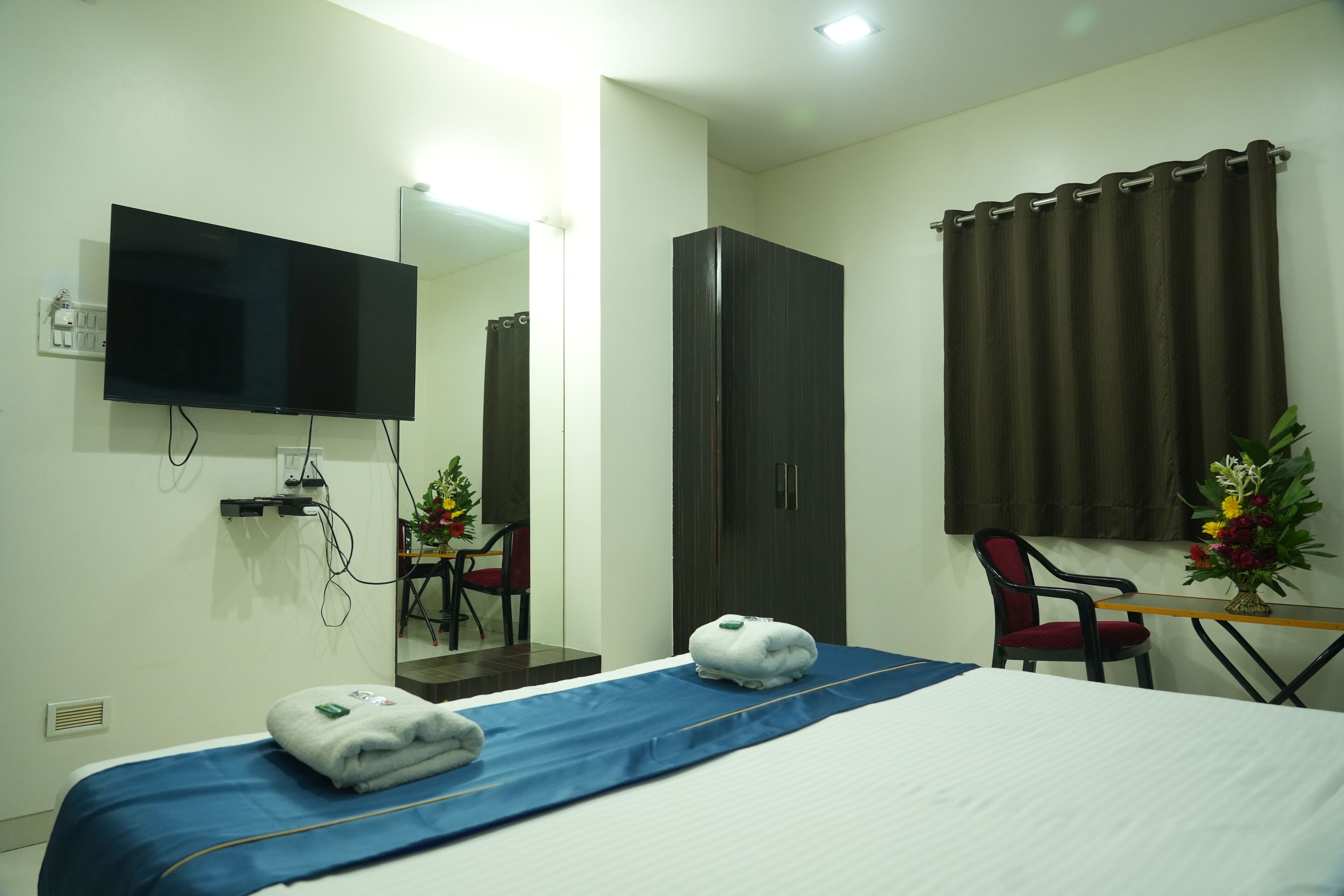 A hotel room with a bed, a flat-screen TV mounted on the wall, a wardrobe, a desk with a chair, and a small table with a vase of flowers.