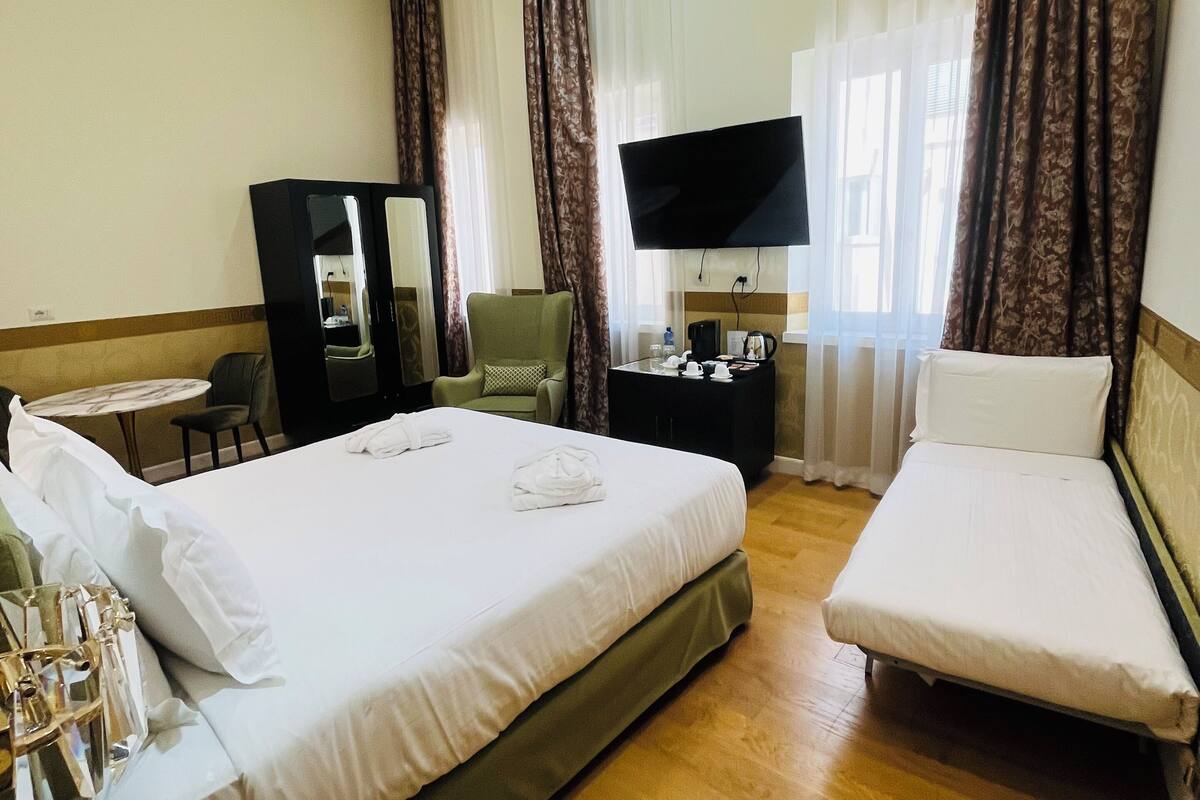 Habitación de hotel con una cama grande, una cama más pequeña, un escritorio con silla, televisión y espejo.