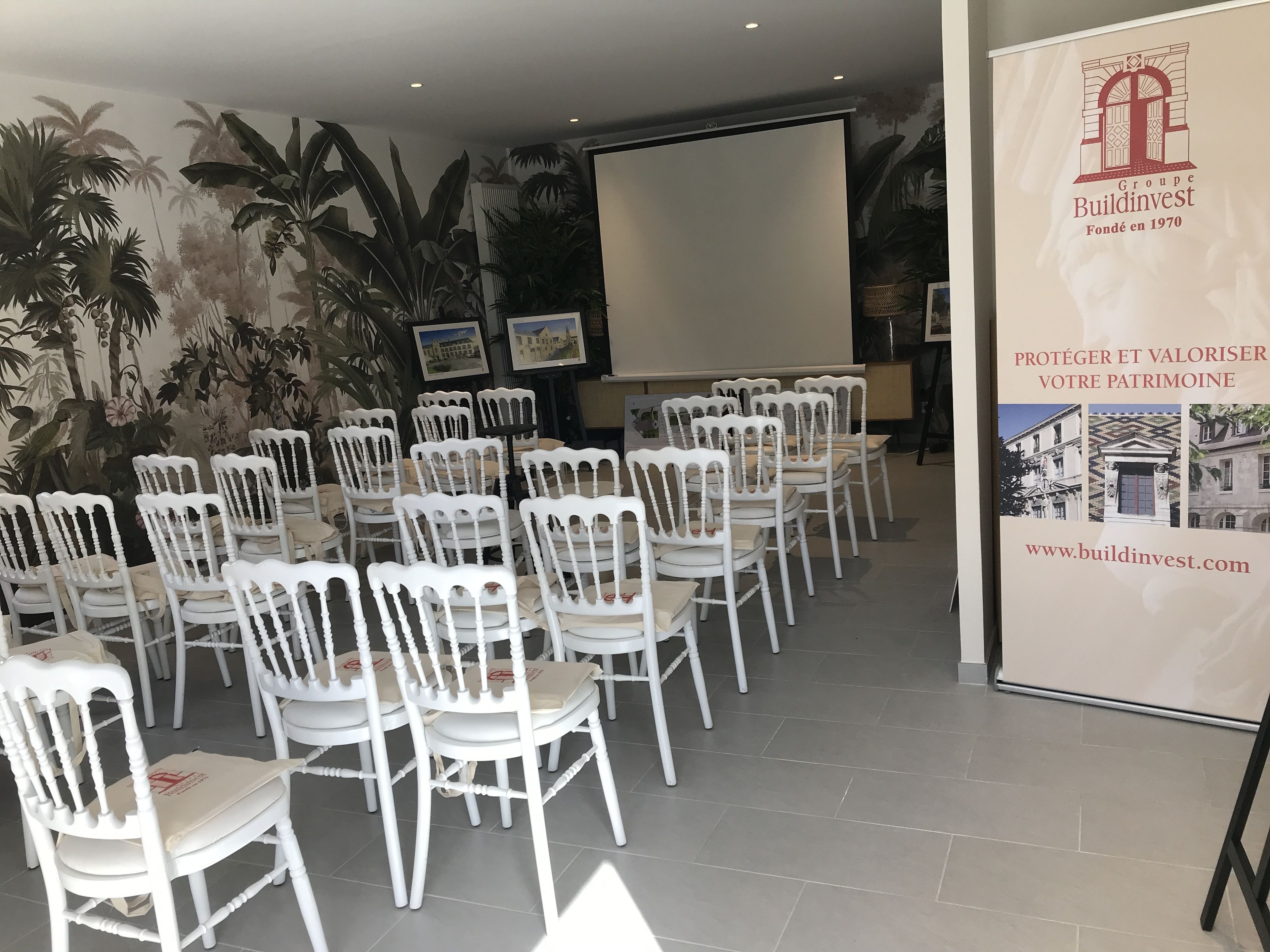 Une salle de conférence avec des chaises blanches disposées en rangées, un écran de projection et une banderole ornée du logo de BuildInvest.
