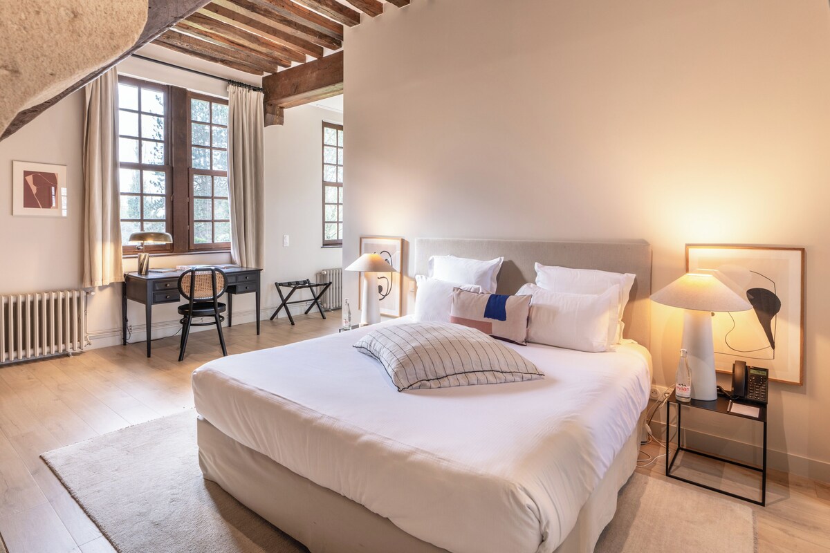 Habitación de hotel con una cama grande, un escritorio y una silla. Hay dos ventanas con cortinas, un radiador y una lámpara en la mesita de noche.