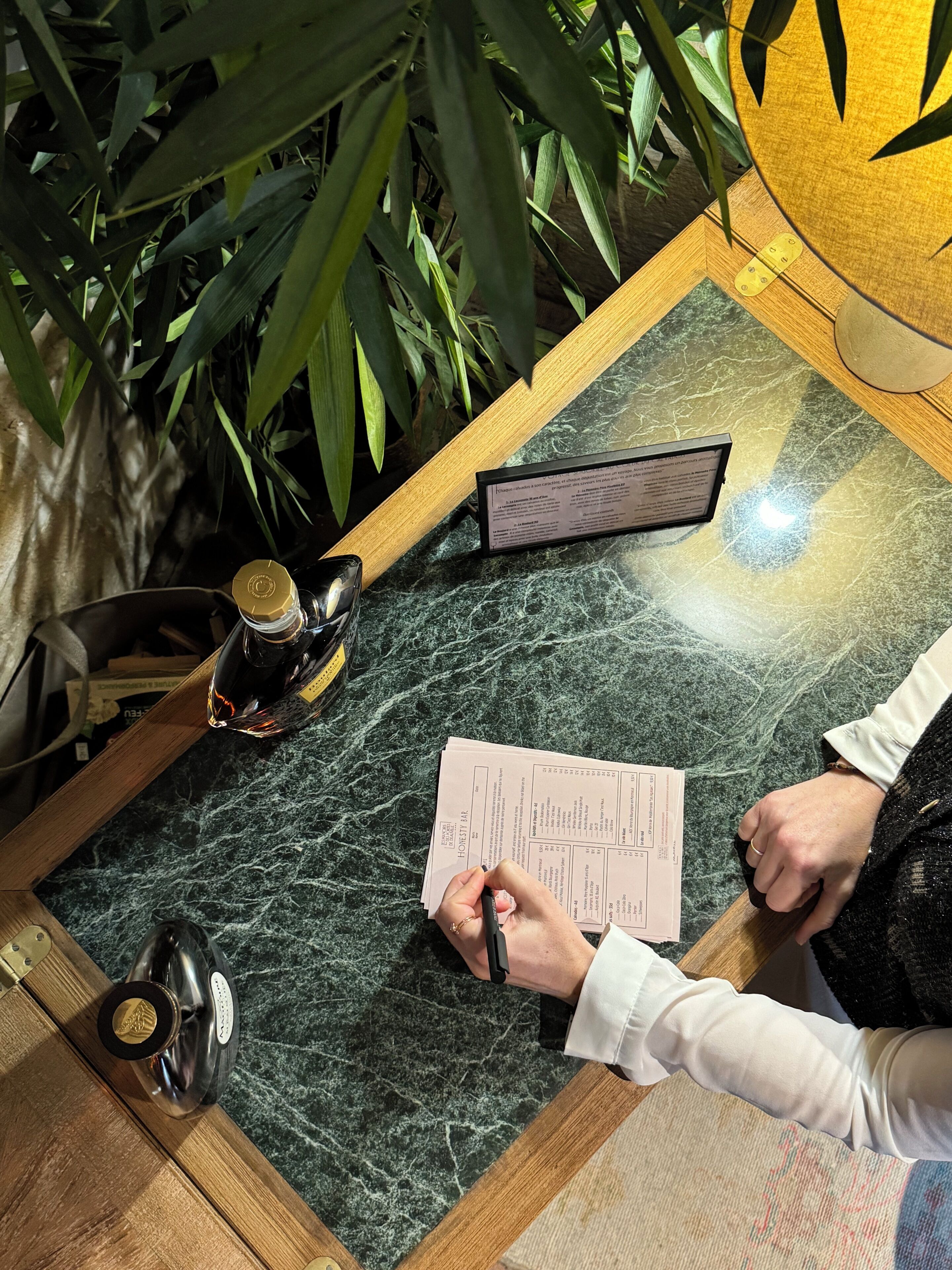 Uma pessoa escrevendo em um papel sobre uma mesa, com um cardápio, uma garrafa e uma planta ao fundo.