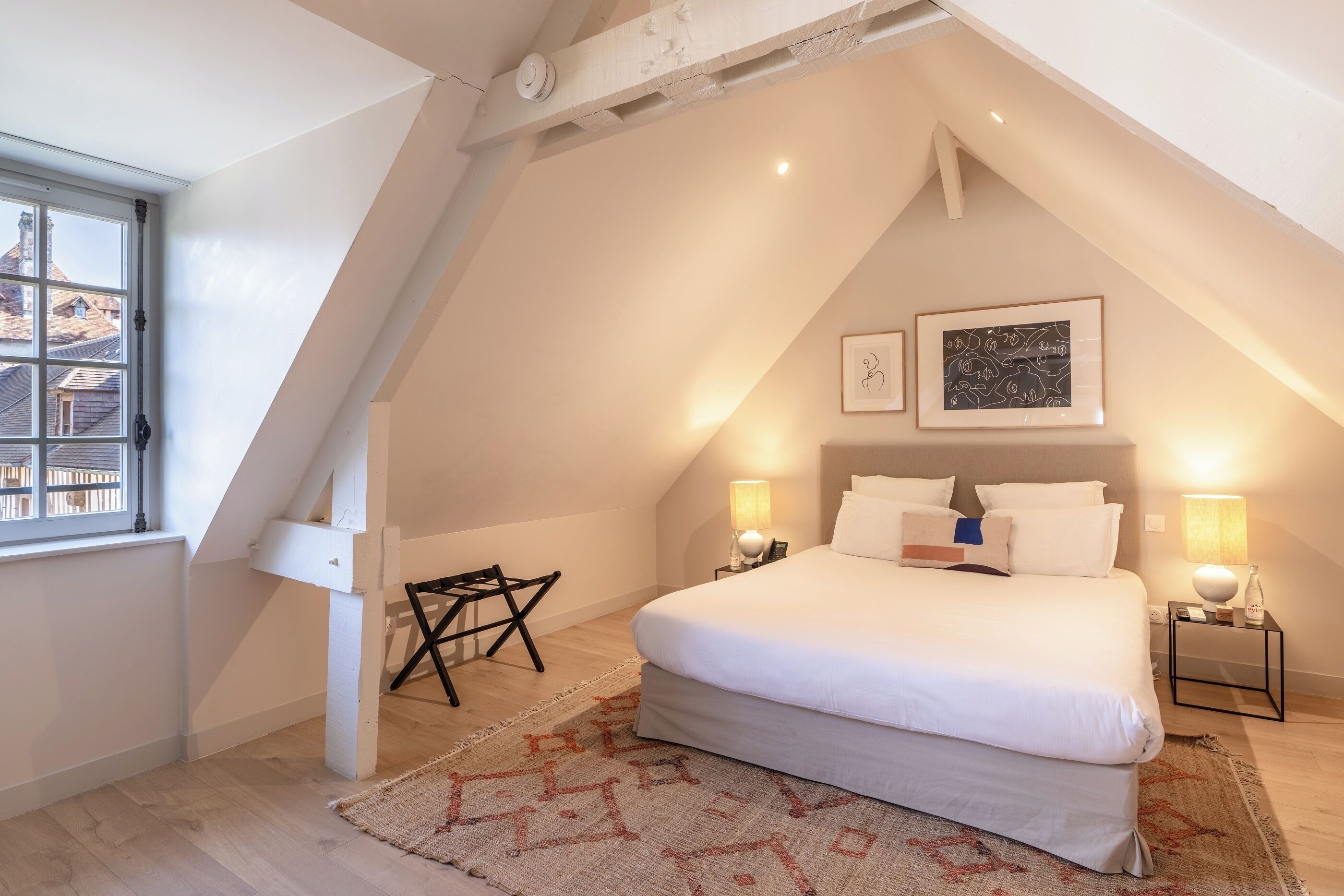 Un dormitorio con una cama grande, dos lámparas de noche, una mesita, una silla y una ventana con vistas a edificios.