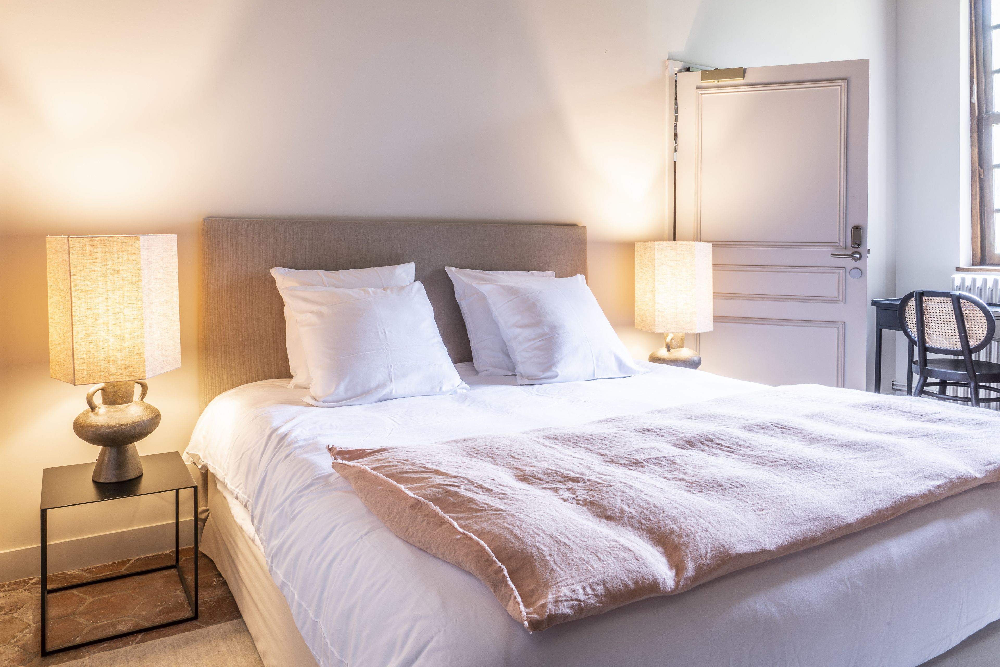 Un lit bien fait, avec du linge de lit blanc et une couverture moelleuse, flanqué de deux lampes de chevet aux abat-jour beiges.