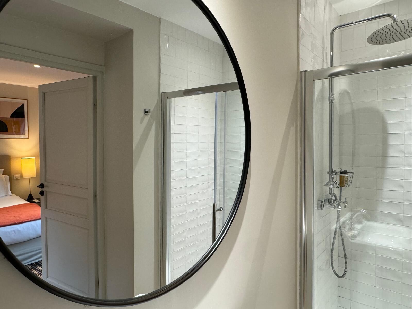 A hotel bathroom with a glass shower enclosure, a round mirror, and a bed with a red accent in the adjacent room.