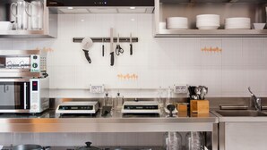 A modern kitchen with stainless steel appliances, including a microwave, espresso machine, and a coffee maker. There are utensils hanging on the wall and a variety of kitchenware on shelves. - Lobby Collective Hostel (Lecce)