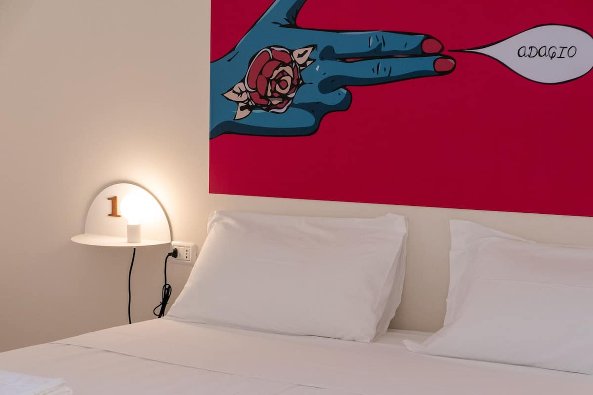 A hotel room with a bed, white pillows, a nightstand with a lamp, and a wall with a graphic of a hand holding a rose and the word "adagio".