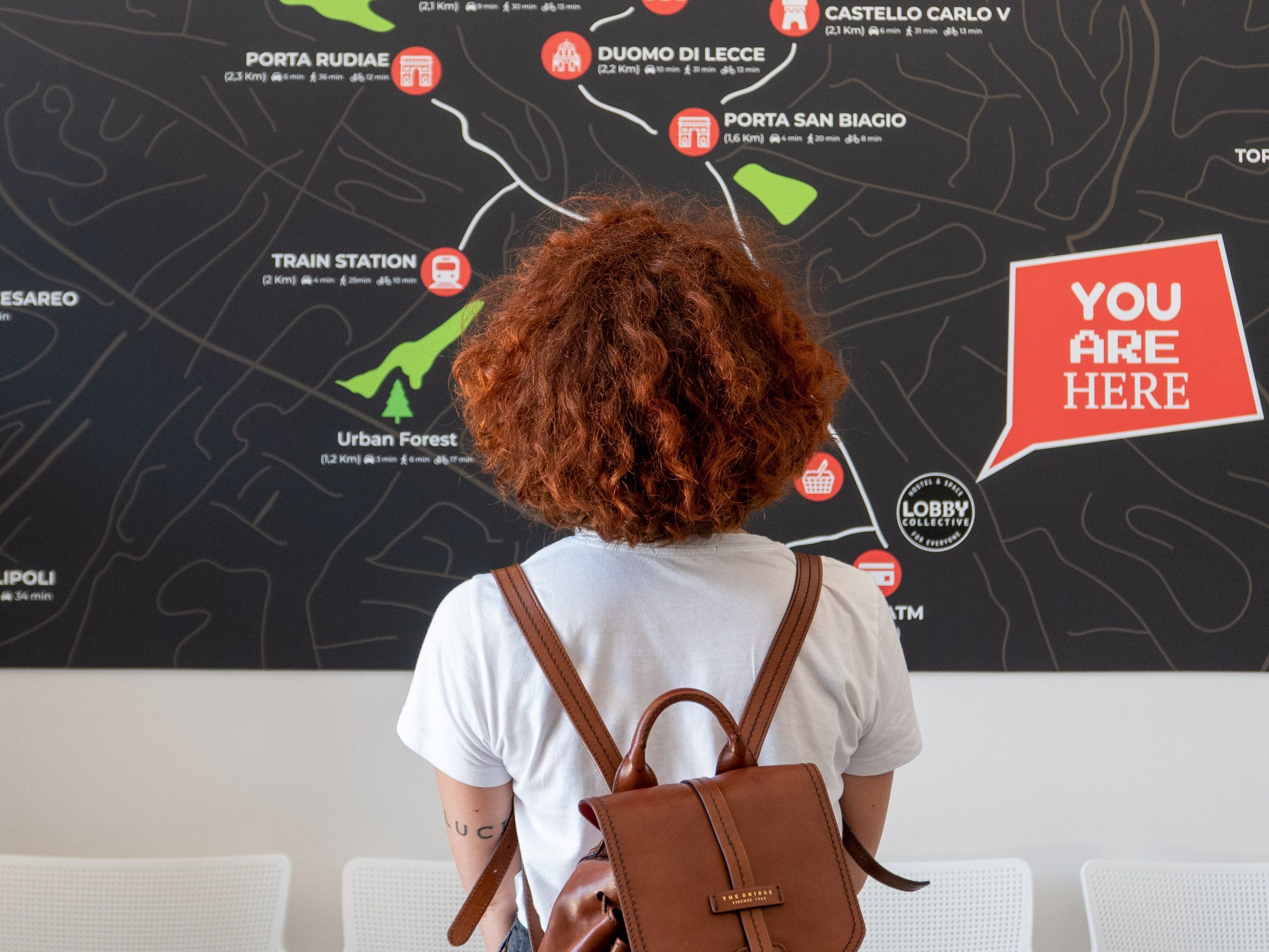 Una persona con una mochila marrón parada frente a un mapa con un bocadillo de diálogo que dice 