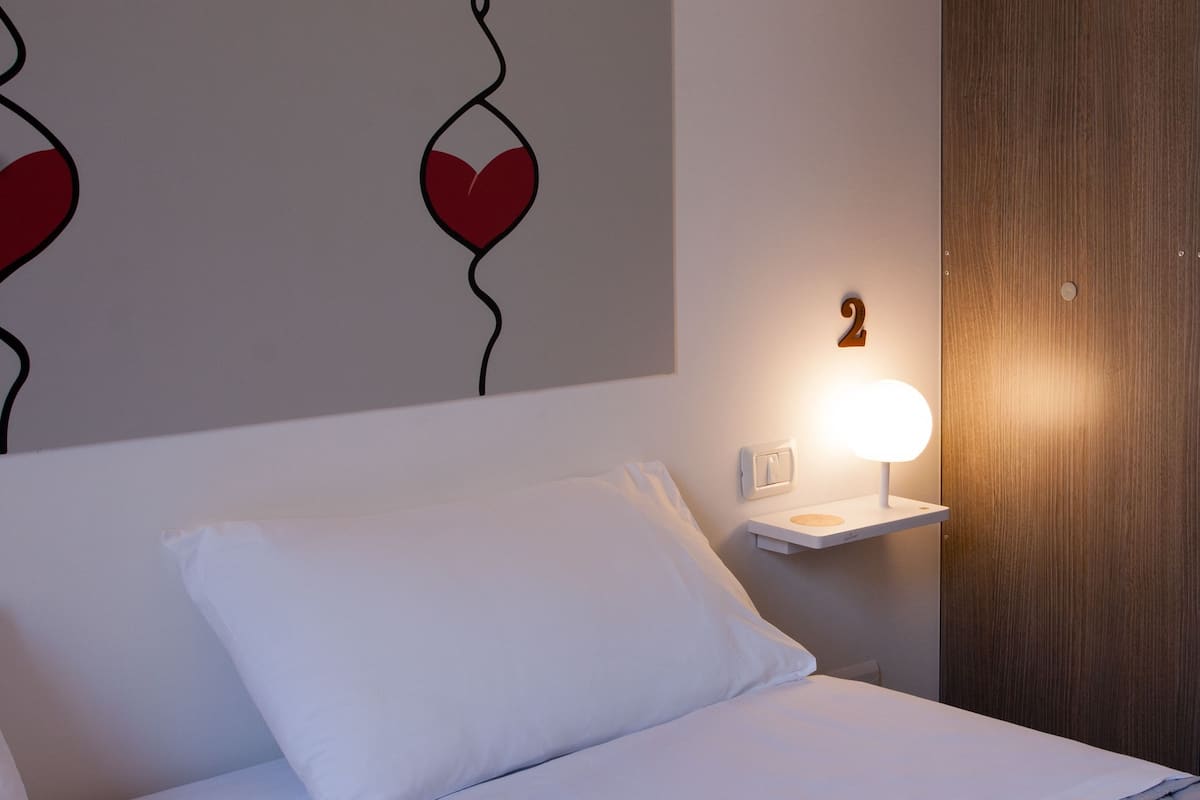 A neatly made bed with white linens, a bedside table with a lamp, and a wall with a number '2' and heart-shaped designs.