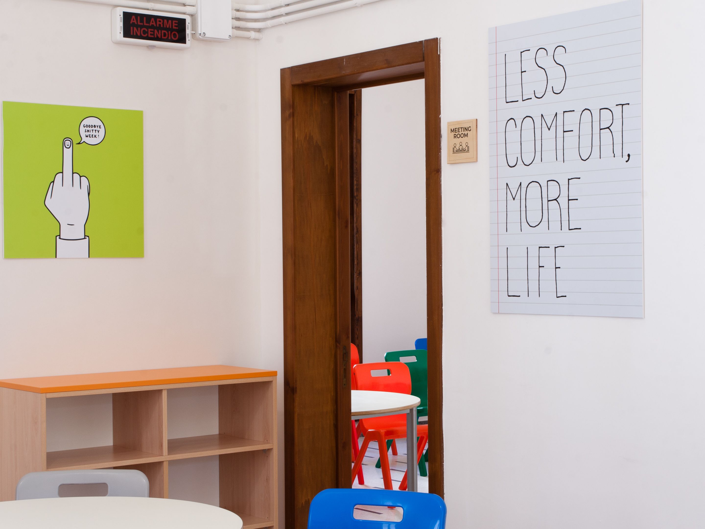Ein Zimmer mit Holztür, weißem Tisch, blauem Stuhl und einem Schild mit der Aufschrift 'LESS COMFORT, MORE LIFE'.