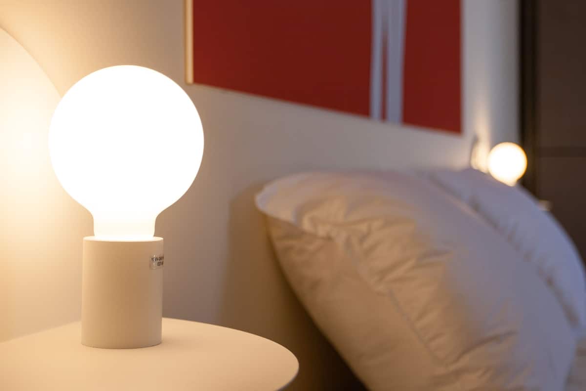 A bedside table with a round, illuminated lamp, a white pillow, and a red wall in the background.