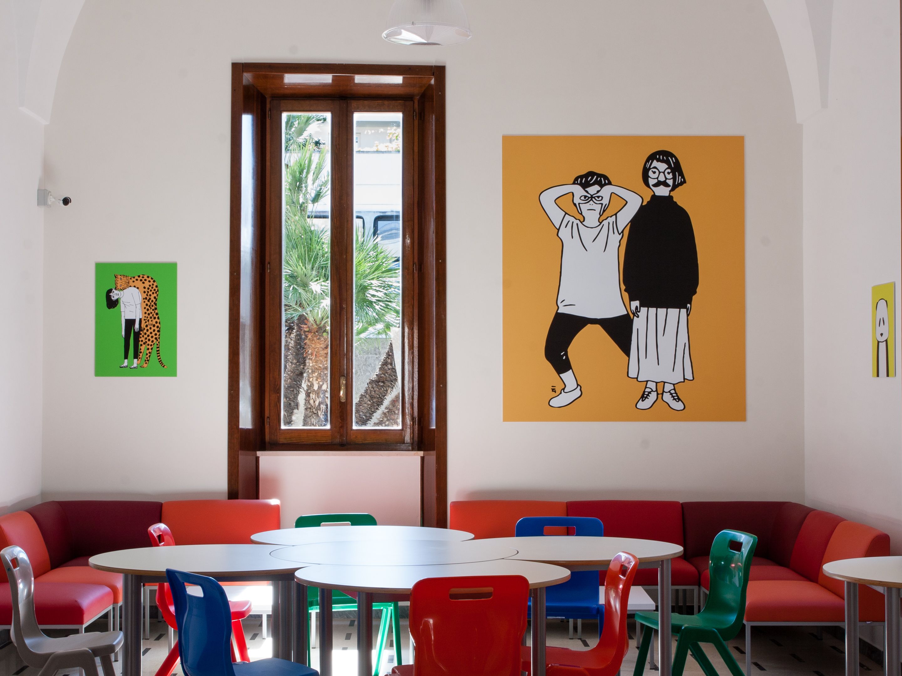 A dining area with colorful chairs and tables, a large window with a view of palm trees, and two wall art pieces.