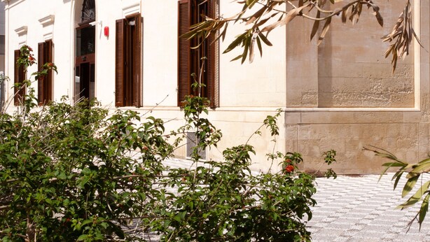 A building with white walls and brown shutters, a cobblestone courtyard, and green foliage. - Lobby Collective Hostel (Lecce)