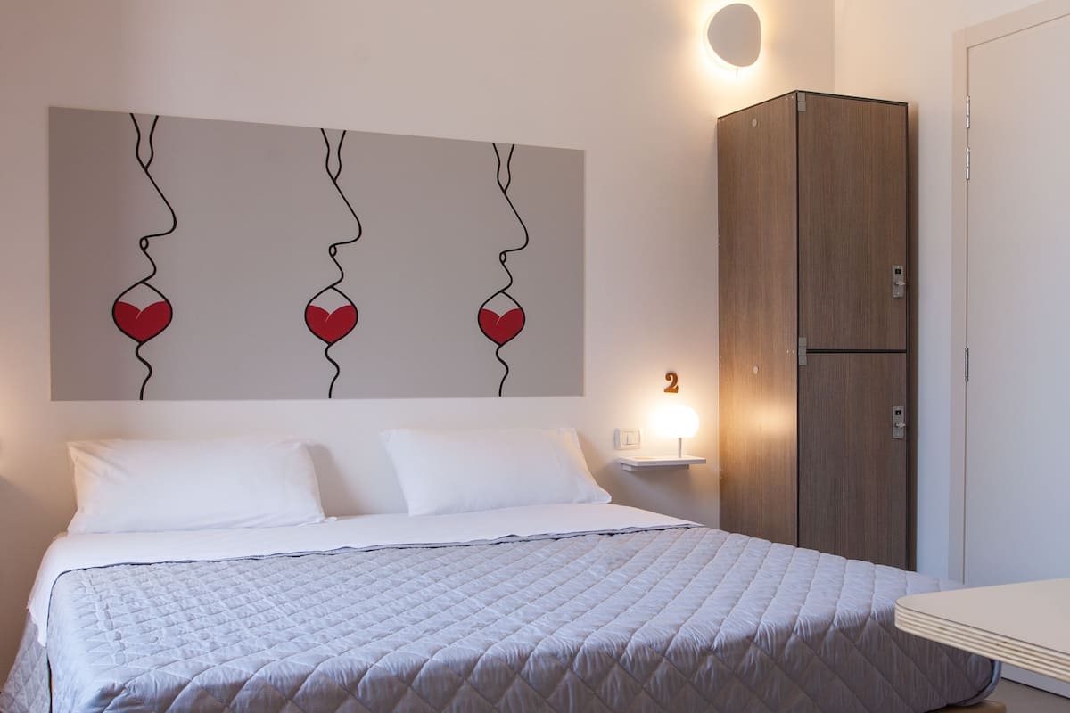 A neatly made bed with a quilted white duvet, a wooden wardrobe, and a wall-mounted light fixture.