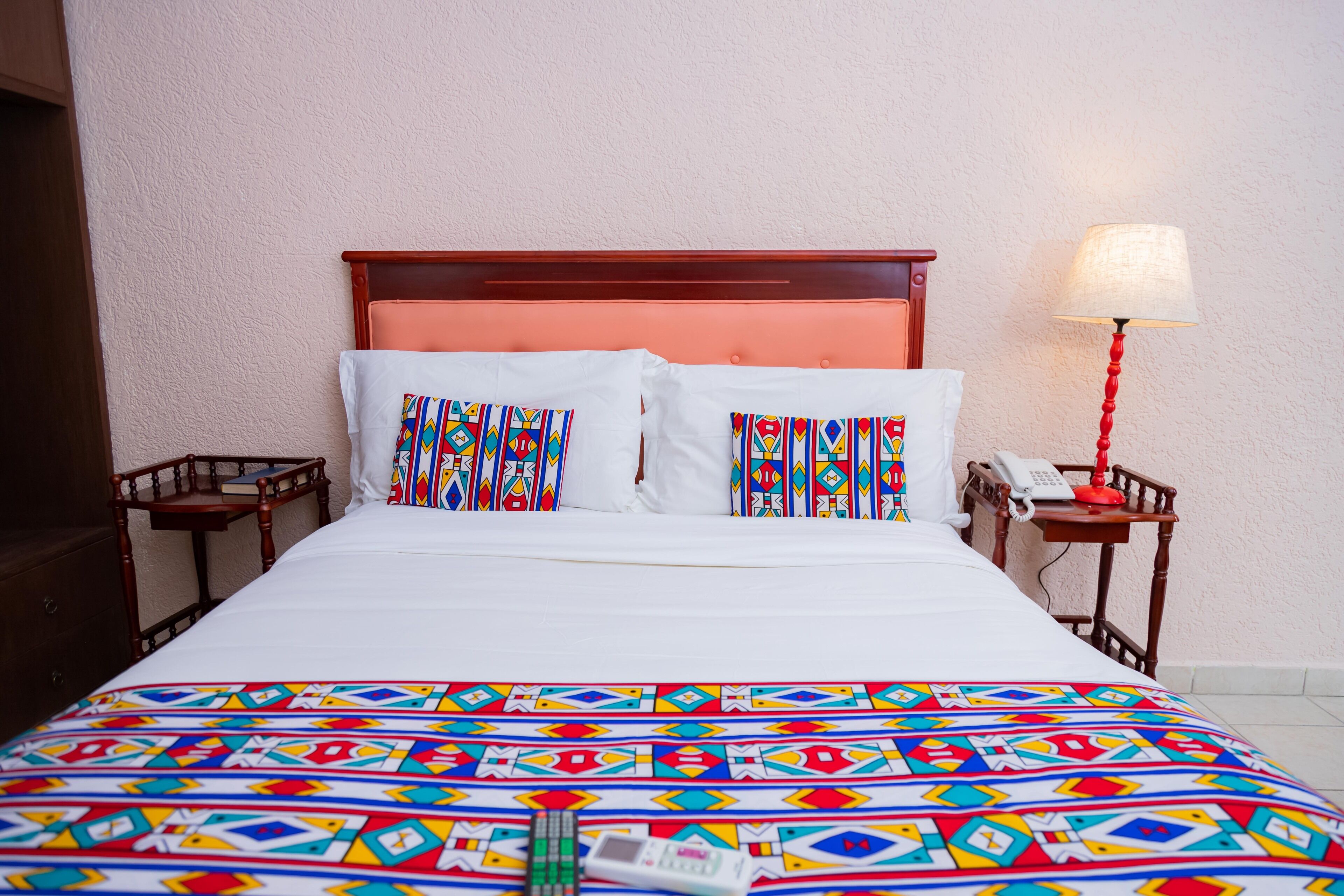A bed with a colorful patterned bedspread, two pillows with a similar design, a wooden headboard, and a bedside table with a lamp and a phone.