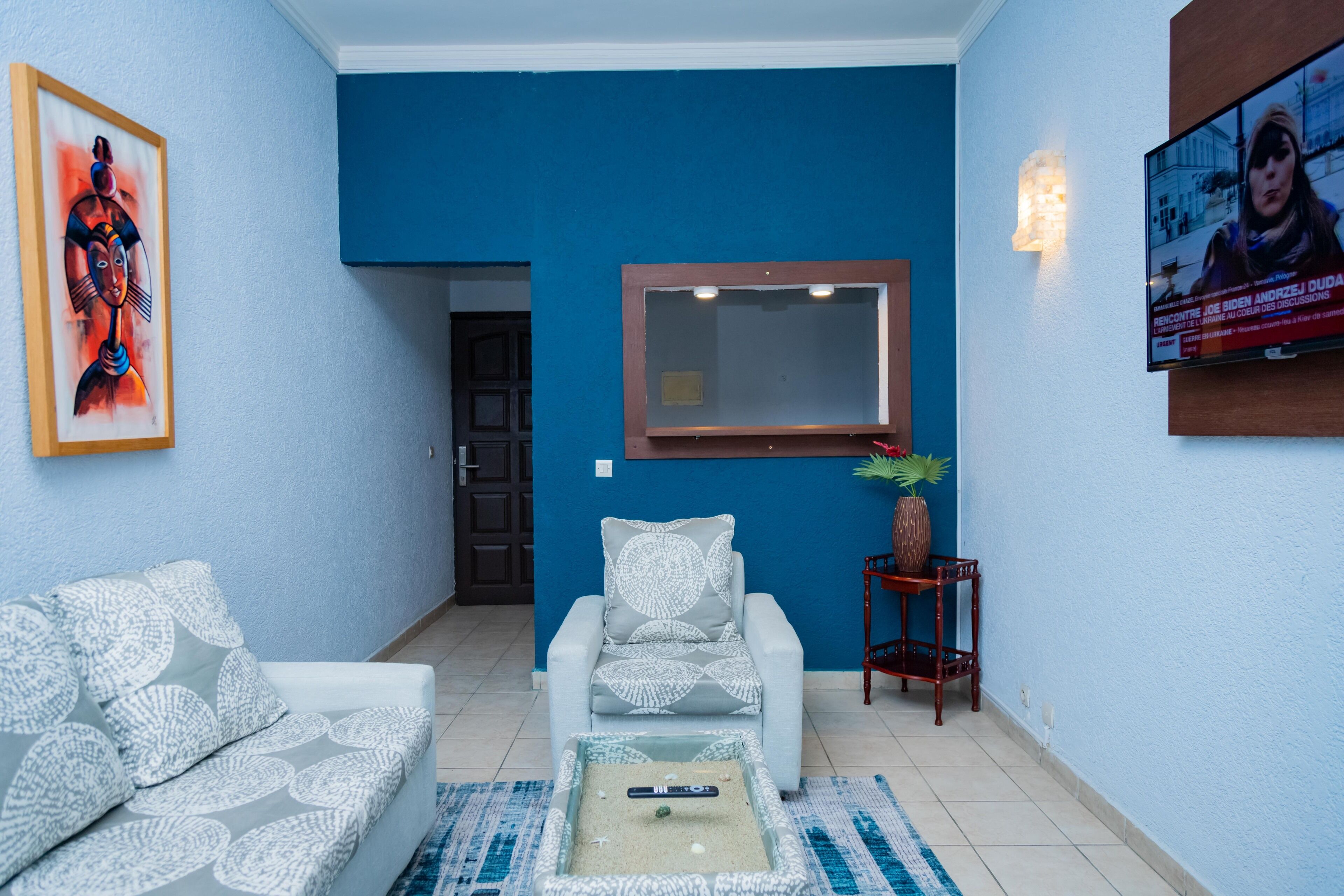 A living room with a blue wall, a white sofa, a patterned armchair, a small table with a remote control, and a television mounted above the armchair.