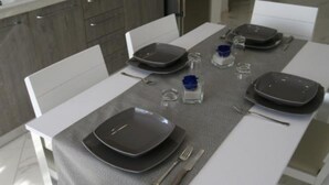 Un juego de mesa moderno dispuesto con platos, cubiertos y copas en una habitación luminosa, con una cocina al fondo.