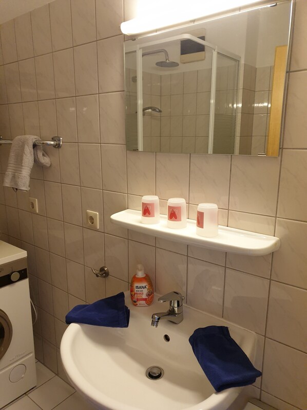A bathroom with a sink, mirror, and shower. There are toiletries on the shelf and a bottle of shampoo.