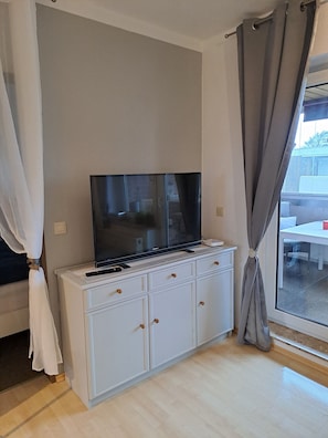Una stanza con una televisione a schermo piatto su una base bianca, una finestra con tende e vista all'esterno.