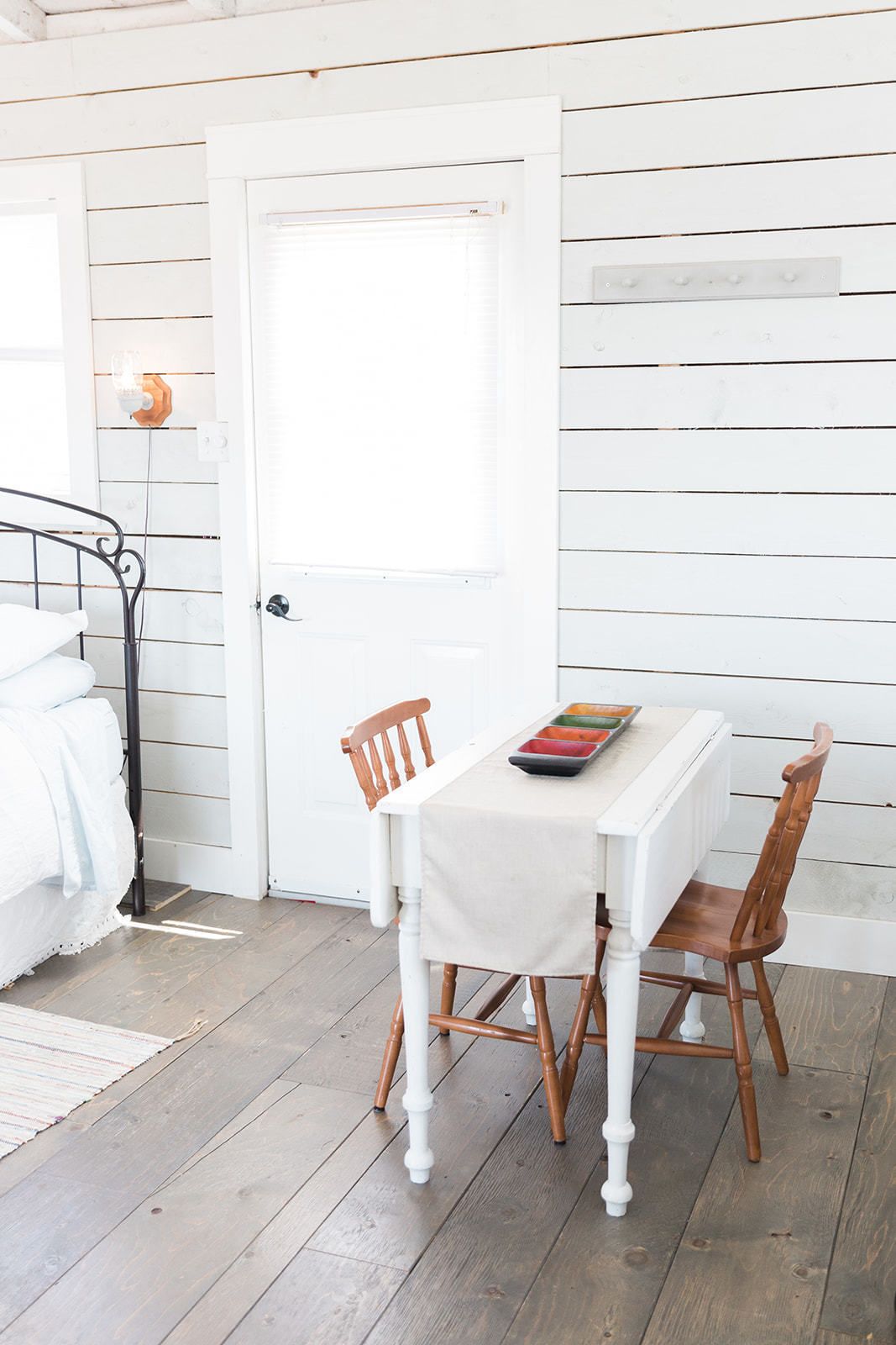 Une pièce avec une porte blanche, un lit avec du linge de lit blanc, une table recouverte d’une nappe blanche et deux chaises en bois.
