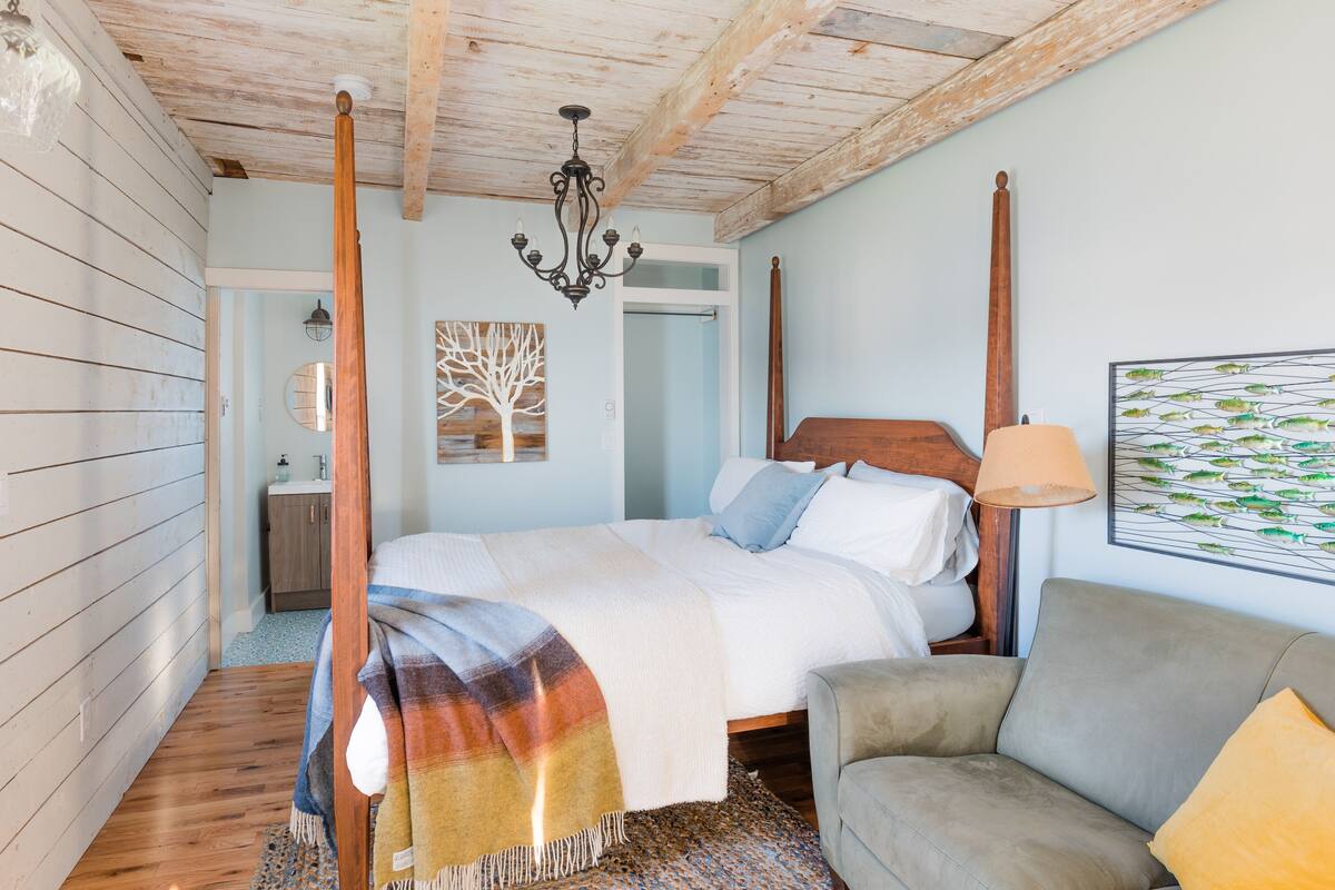 A bedroom with a four-poster bed, a wooden ceiling, a chandelier, a painting of fish, a sofa, and a patterned rug.
