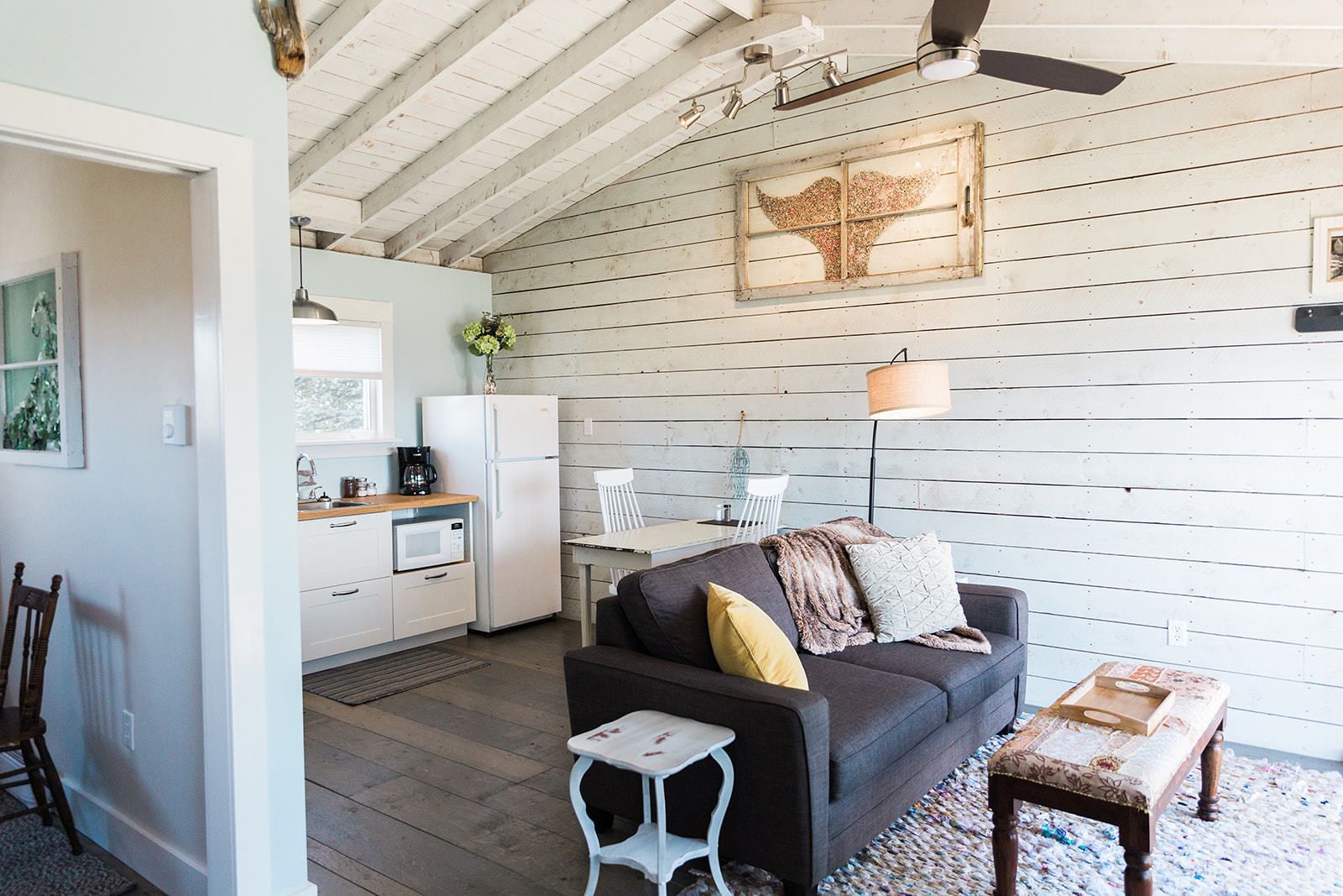 Una sala de estar acogedora con un sofá oscuro, una pequeña mesa blanca y un banco. Se ve, al fondo, una cocina con electrodomésticos blancos y una zona de comedor con una silla y una mesa blancos.