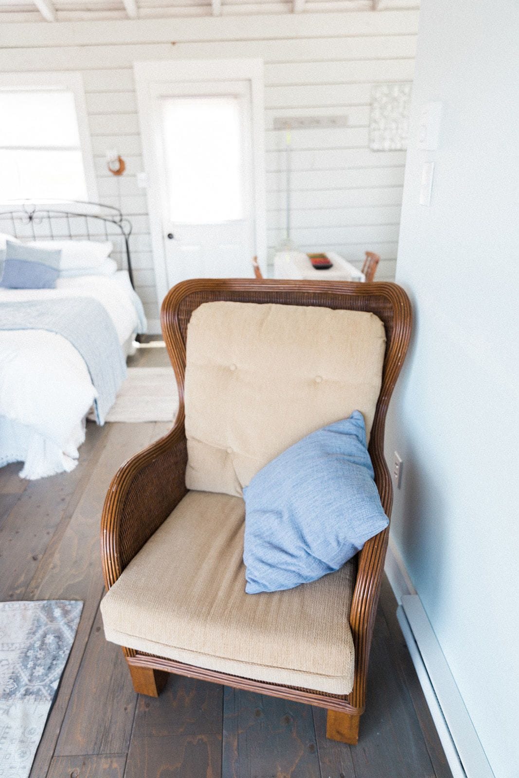 A wicker chair with a beige cushion and a blue pillow in a room with a bed, a white door, and a patterned rug.