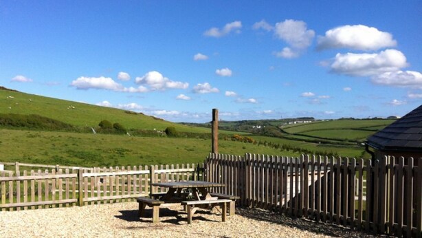 A picnic area with a wooden table and benches, surrounded by a wooden fence, set against a backdrop of rolling green hills and a clear blue sky. - Merlin Farm Daymer and Treyarnon (Newquay)