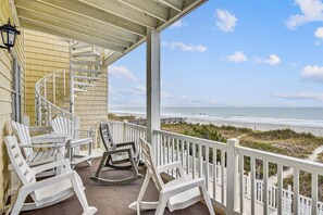 A covered porch with white rocking chairs and a spiral staircase leading to a balcony overlooking a beach.