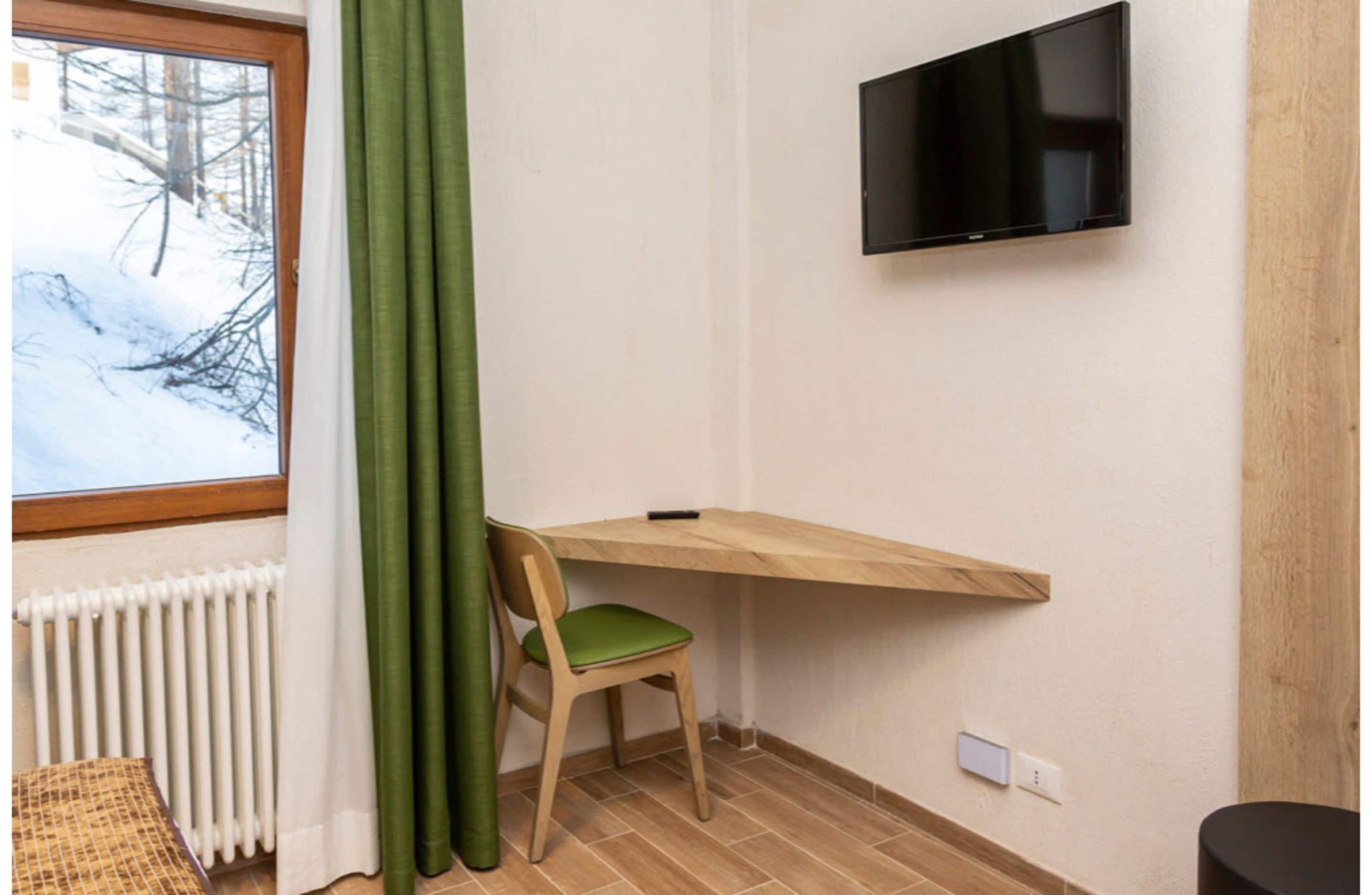 A room with a wooden desk, a chair, a television mounted on the wall, and a window with a view of trees and snow.
