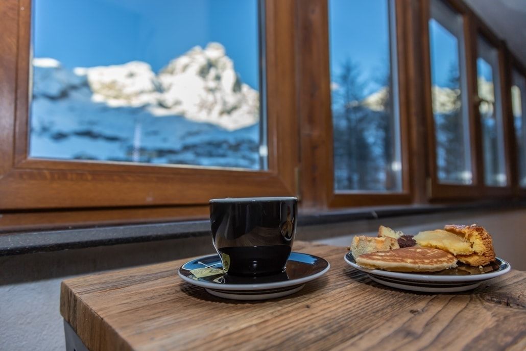 A wooden table with a black cup and saucer, and a plate of pancakes and pastries by a window with a view of rocks and trees.
