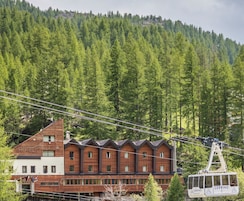 Un hotel con fachada de madera y numerosas ventanas, un teleférico en primer plano y una montaña boscosa al fondo.
